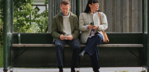 Par sitter vid busshållplats med sina mobiltelefoner i handen. Mannen till vänster tittar ner i sin telefon.
