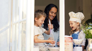 Vuxen och två barn syns genom fönster, bakar i kök