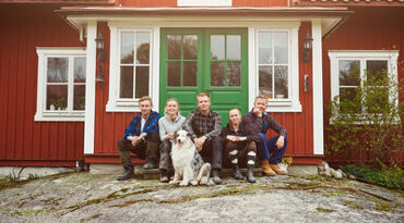Fem personer och en hund sitter på en trapp framför en grön dörr och ett rött hus