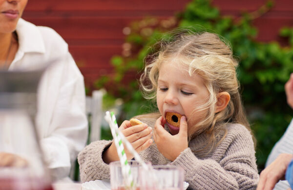 Flicka äter en kaka utomhus vid ett bord. Saftglas med sugrör i förgrunden.