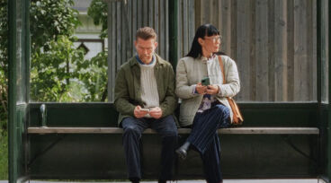 Man och kvinna sitter vid hållplats och väntar på bussen. Mannen tittar ner i sin mobiltelefon.