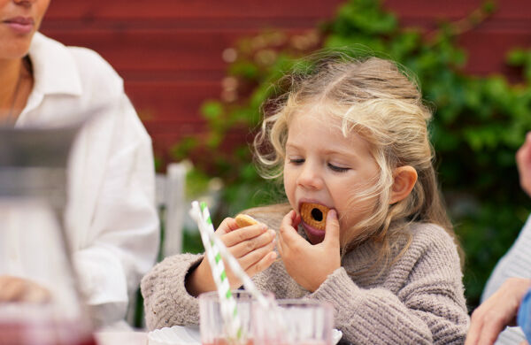Flicka äter en kaka utomhus vid ett bord. Saftglas med sugrör i förgrunden.